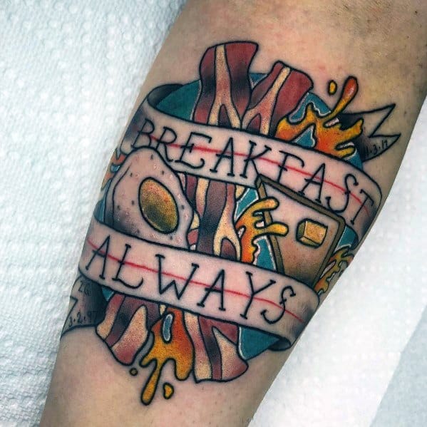Bold traditional-style tattoo featuring bacon, eggs, toast, and melted butter with a banner reading "Breakfast Always," celebrating morning meals.