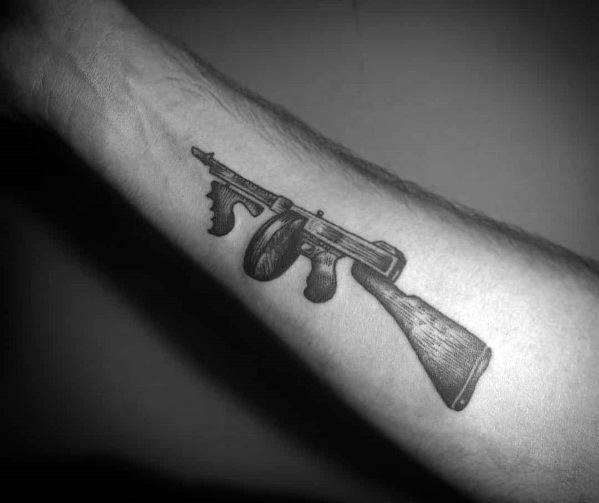 Realistic black and grey Tommy gun tattoo on the forearm, featuring fine shading and intricate details for a classic and bold firearm design.