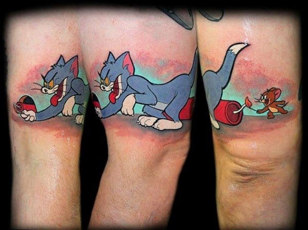 Colorful thigh tattoos of Tom holding a lit firecracker and Jerry running away with the fuse, stretched across two legs in cartoon style.