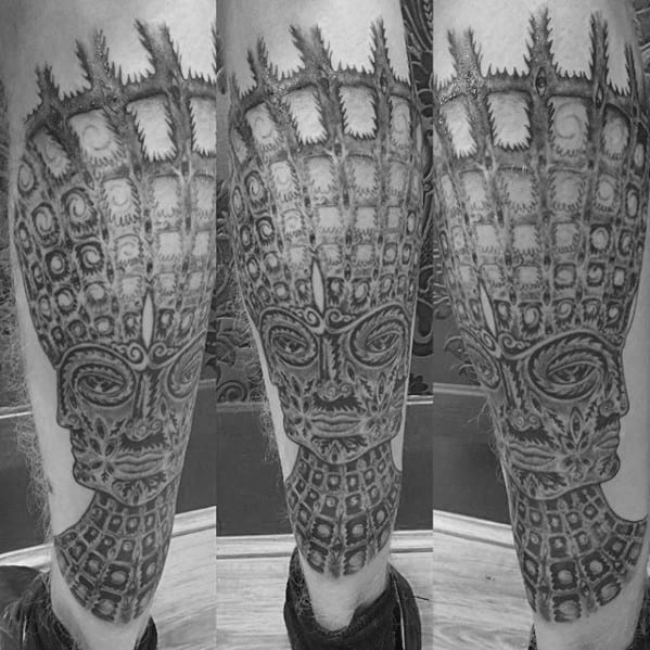Black and grey Tool-inspired leg tattoo of a symmetrical, alien-like face with grid patterns, ornate details, and vertical eye motifs.