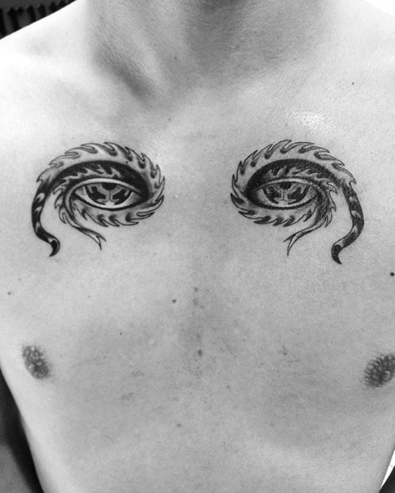 Black and grey Tool-inspired chest tattoos of two symmetrical eyes with tribal flame designs, placed above each pectoral muscle.