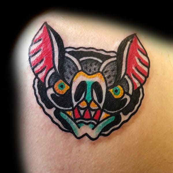 Colorful traditional bat head tattoo with bold black outlines, red ears, teal accents, and vibrant yellow and green eyes.