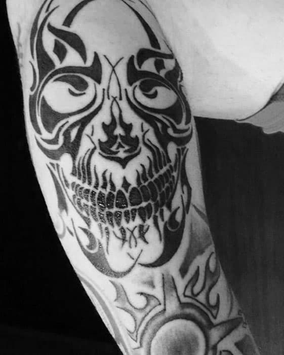 Tribal skull tattoo on upper arm with sharp black patterns, intense eyes, and detailed teeth forming a bold symmetrical design.