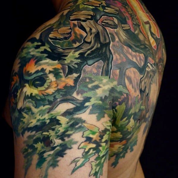 Colorful tattoo of a forest scene with a hidden owl face blending into leaves and a large tree trunk across the shoulder and back.