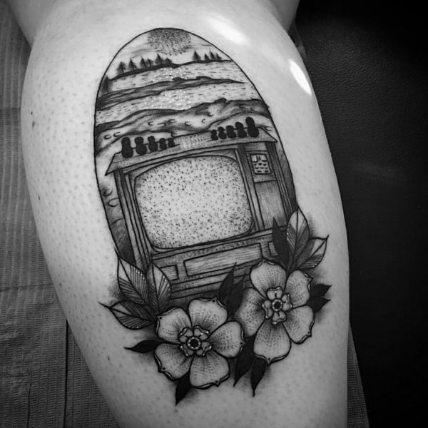 Black and grey tattoo of a vintage TV with a static screen, framed by flowers, with an outdoor landscape emerging from the top, blending nature and nostalgia.