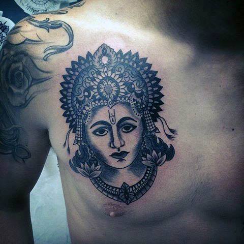 Black ink Krishna tattoo on the chest showing a bold, symmetrical face adorned with an ornate crown, lotus motifs, and spiritual markings.