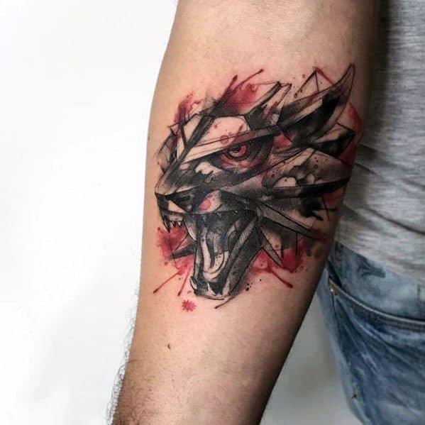 Watercolor-style Witcher tattoo on the upper arm featuring the fierce wolf medallion with red splashes and sharp black sketch lines.