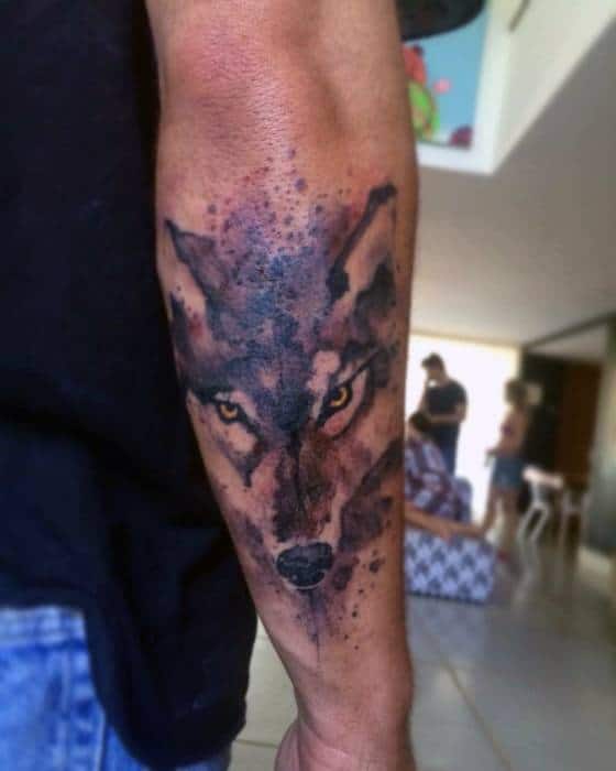 Forearm wolf tattoo with intense yellow eyes, dark watercolor splashes, and a fierce expression blending realism and abstract style.
