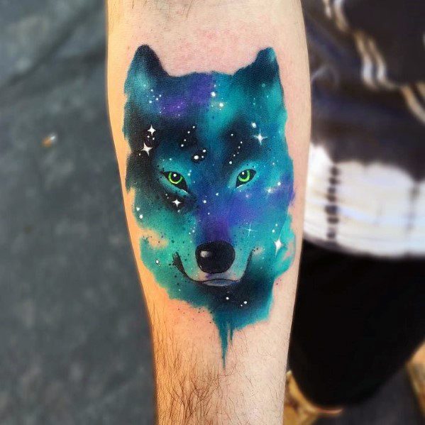 Forearm wolf tattoo with glowing green eyes and a galaxy-inspired design in blue, teal, and purple, accented with white stars.