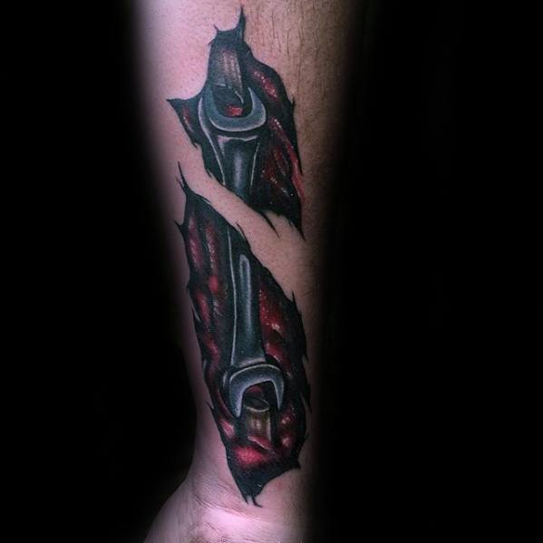 Tattoo of a wrench appearing beneath torn skin with red and black shading, creating a dramatic 3D biomechanical effect on the forearm.