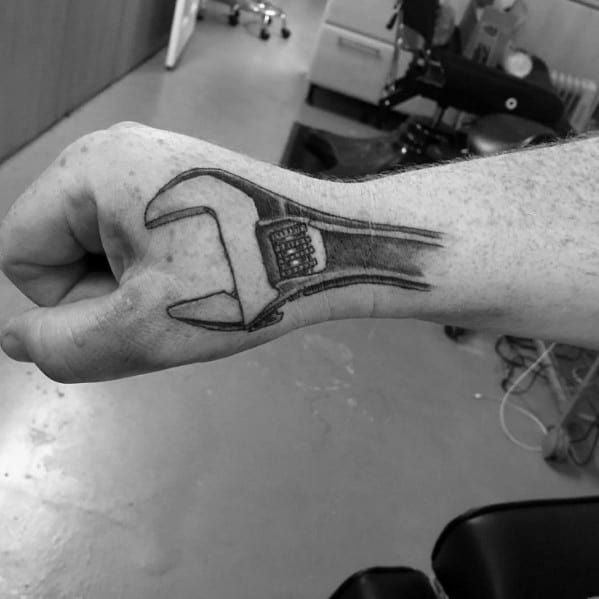 Black ink tattoo of an adjustable wrench extending across the top of the hand and wrist, designed to follow the natural contours.