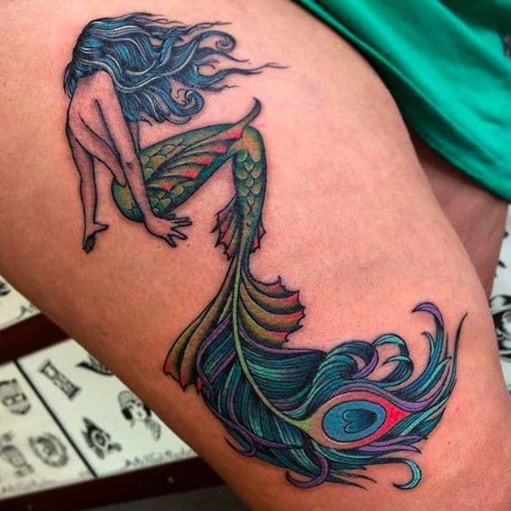 Vibrant thigh tattoo of a mermaid with flowing blue hair and a peacock feather-inspired tail, featuring rich green, blue, and purple hues with intricate details.
