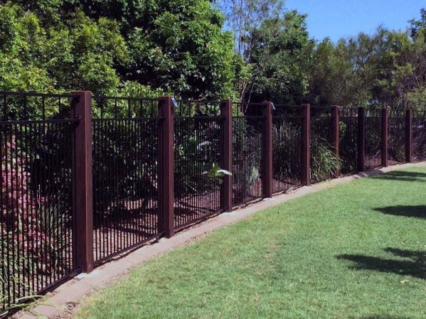 A tall fence with wooden posts borders a lush garden and neatly trimmed lawn