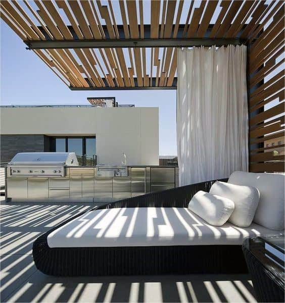 Contemporary pergola with wood slats, outdoor lounge, white curtains, and built-in kitchen area.