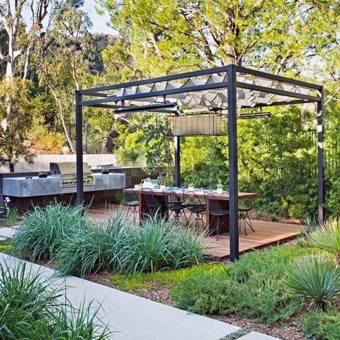 Modern metal pergola pavilion with outdoor dining area and built-in grill, surrounded by lush greenery.