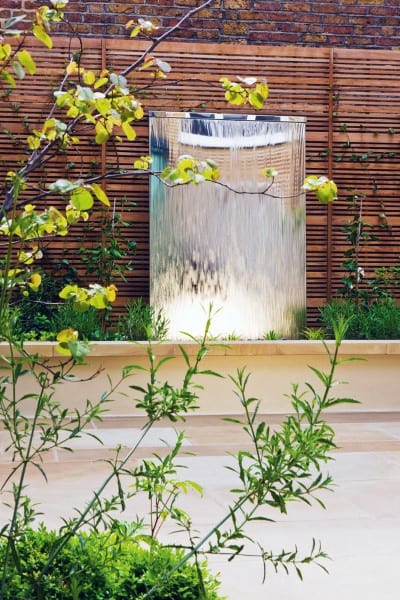 Sleek stainless steel waterfall feature with bamboo fence and lush greenery in a modern garden.