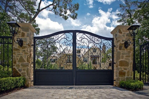 Black wrought iron driveway gate with elegant scrollwork and stone pillar accents.