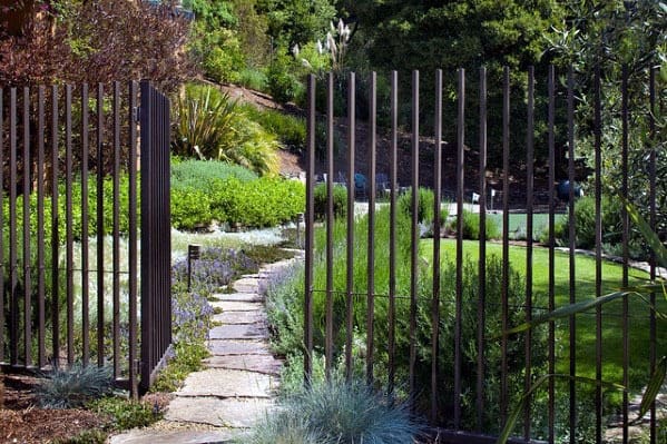 A garden with a stone path, fence, and metal gate leading to a lush lawn and trees in the background