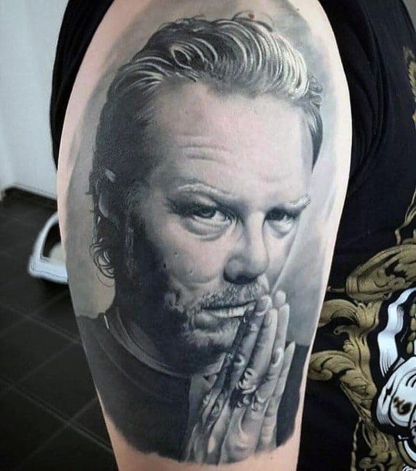 Realistic upper arm portrait tattoo of James Hetfield with hands clasped near his face, detailed in soft black and grey shading.