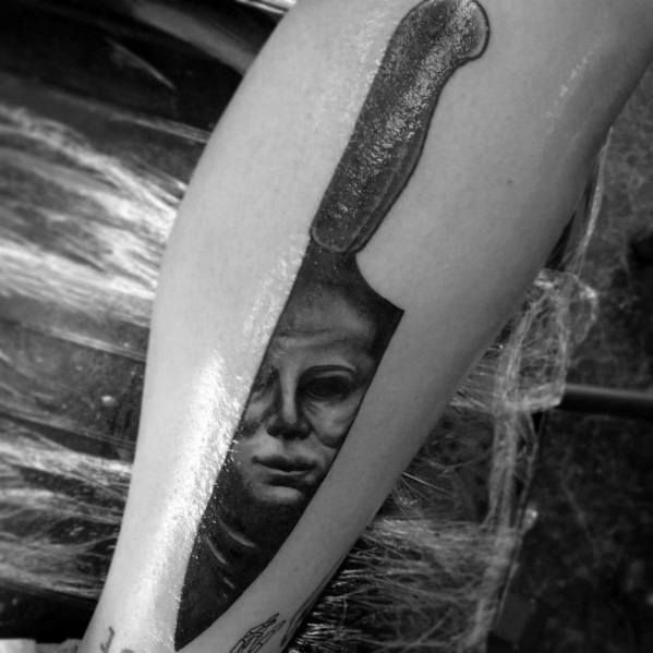 Unique black and gray Michael Myers tattoo, featuring his haunting face reflected in a large, realistic knife blade for a chilling effect.