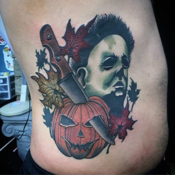 Bold and colorful Michael Myers tattoo featuring his eerie face alongside a sinister jack-o'-lantern, a large knife, and autumn leaves, blending horror and Halloween themes.