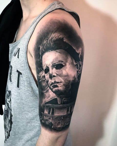 Realistic black and gray Michael Myers tattoo on the upper arm, featuring his ominous mask and the iconic Myers house below.