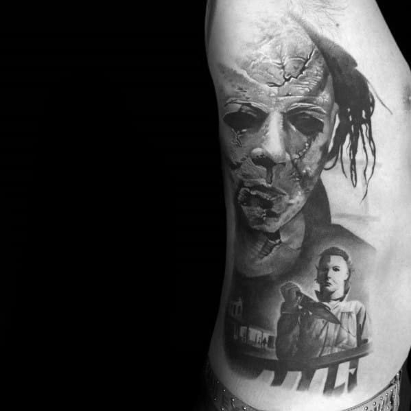 Black and grey Michael Myers tattoo featuring his cracked mask with eerie shading and a haunting scene from Halloween below.