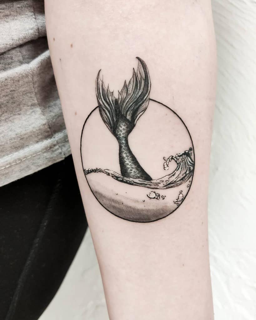 Black and grey tattoo of a mermaid’s tail emerging from water within a circular frame, with detailed shading and wave elements.
