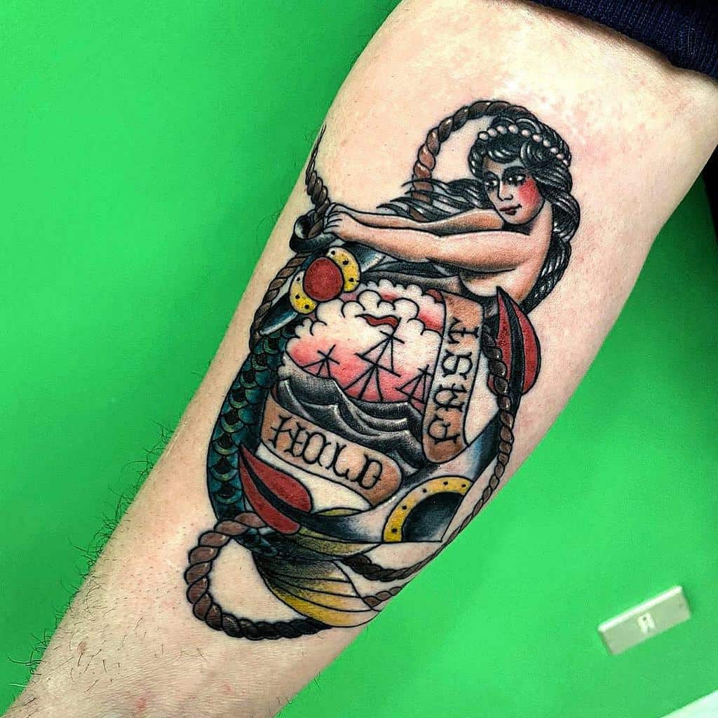 Traditional tattoo of a mermaid wrapped around a ship in a bottle, holding a rope, with 'Hold Fast' text and nautical elements.