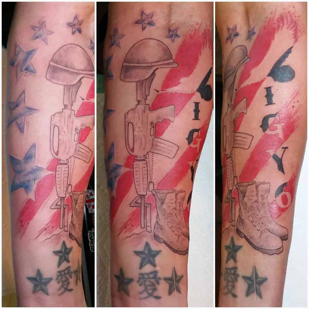 Forearm tattoo featuring a battlefield cross with helmet, rifle, and boots, set against a red-striped American flag with 'IGY6' text