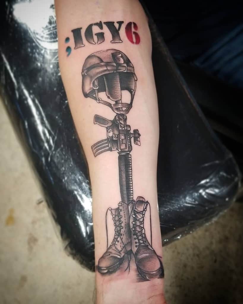 Forearm tattoo of a rifle, helmet, and boots forming a memorial. Above, there's text reading "; IGY6" in black and red