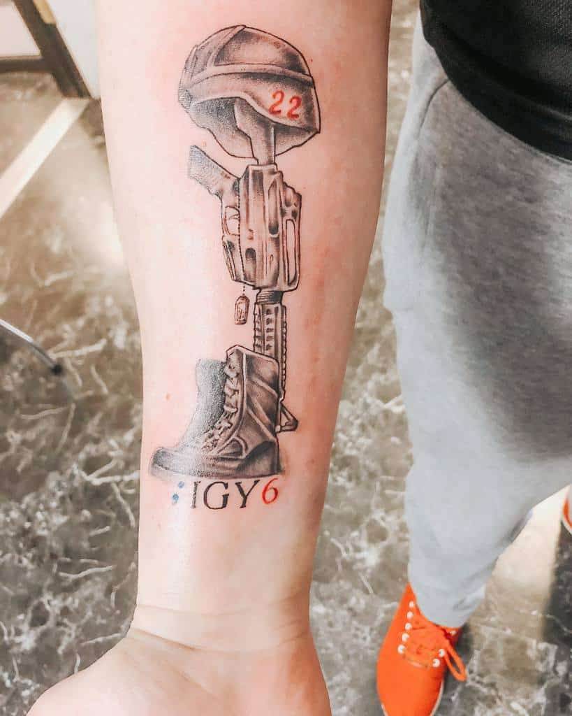 Forearm tattoo of a battlefield cross with helmet, rifle, and boot, featuring 'IGY6' and '22' in red, symbolizing remembrance