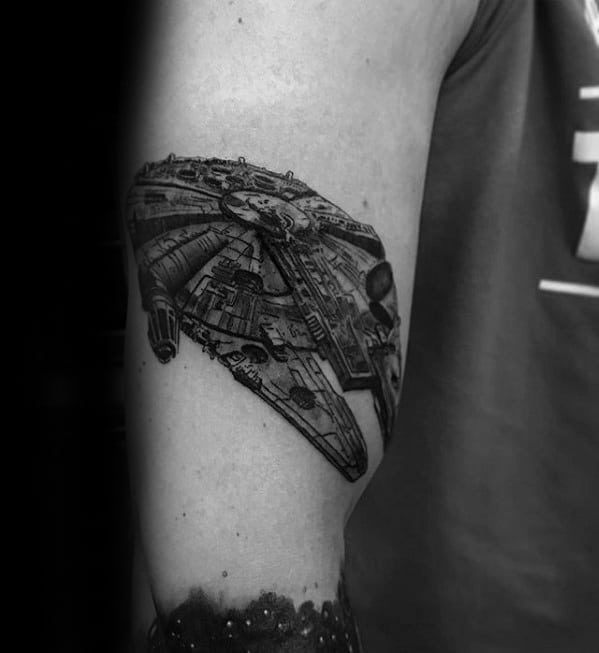 Detailed black and grey Millennium Falcon tattoo on the upper arm, with realistic shading and a 3D flying perspective.