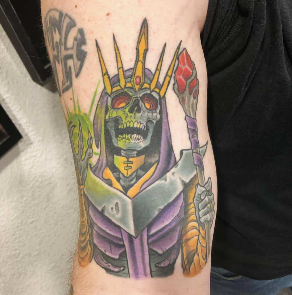 A colorful tattoo of a skeletal lich wearing a golden crown and purple robes, wielding a glowing red staff and a green magical orb