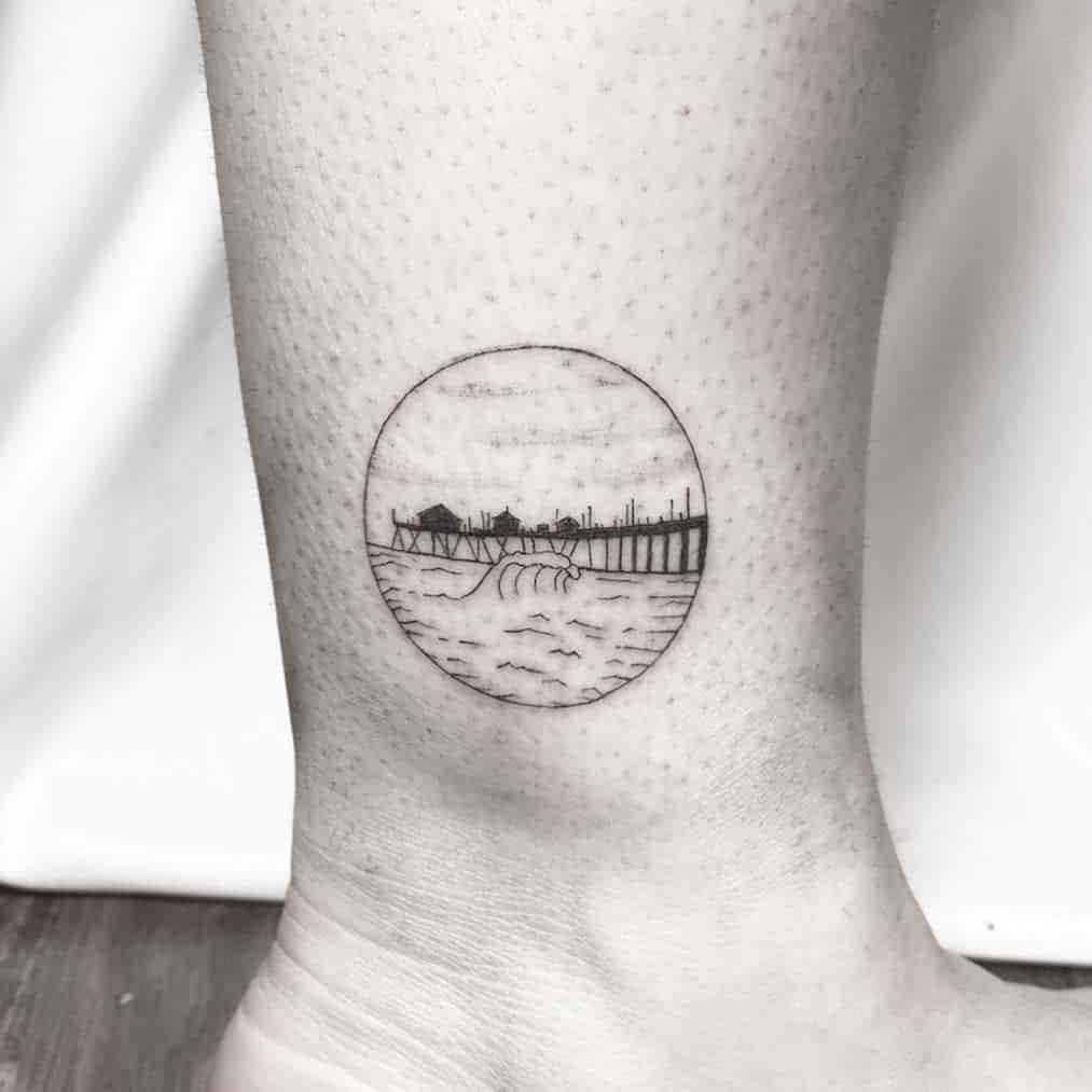 Minimalist tattoo featuring a circular design with an ocean wave, a pier, and small houses in fine line style