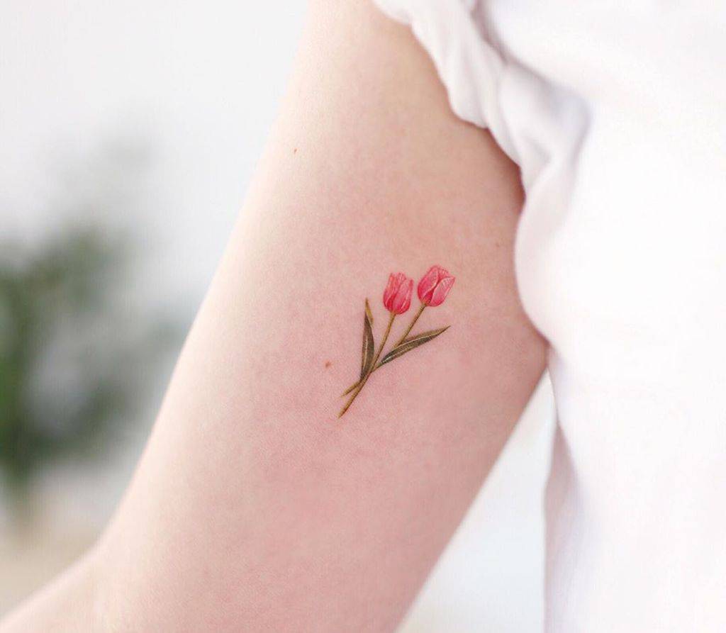 A small, delicate tulip tattoo on the upper arm, featuring two pink flowers with thin green stems and leaves in a fine watercolor style.