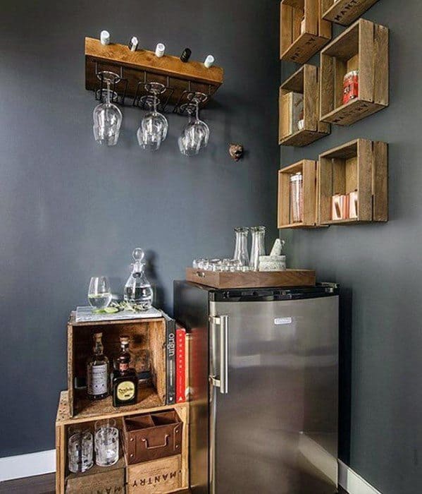 A stylish corner bar with a mini fridge, wooden crates, glasses, and bottles on a dark wall