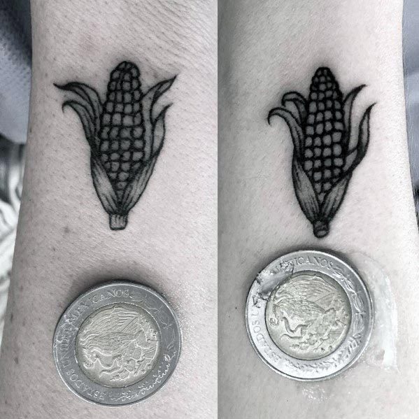 Small black ink tattoo of a corn cob with husks, finely detailed and placed on the wrist, shown next to a coin for scale.