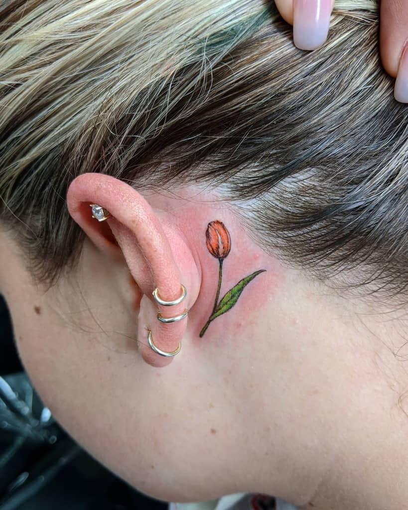 A small, colorful tulip tattoo behind the ear, featuring an orange flower with a green leaf in a delicate, fine-line style.