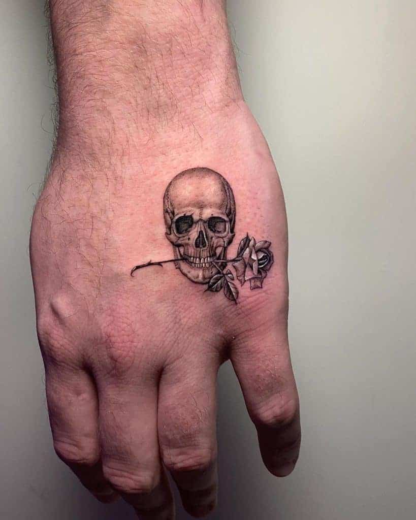 Hand tattoo of a skull holding a rose stem in its teeth
