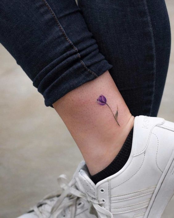 A small, delicate purple tulip tattoo on the ankle, featuring a fine stem and a single green leaf in a minimalist watercolor style.