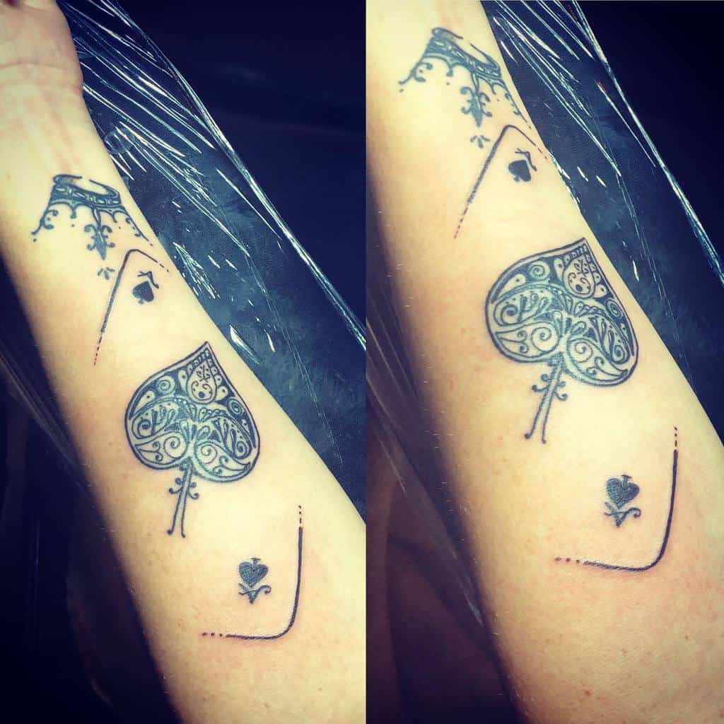 A black-and-white tattoo of the Ace of Spades with ornate detailing, accompanied by minimalistic lines and small spade symbols near the design
