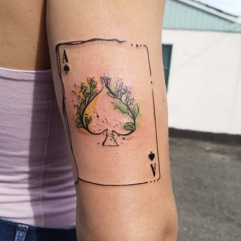 A tattoo of the Ace of Spades card, with the spade symbol formed by a colorful floral design, featuring shades of green and yellow