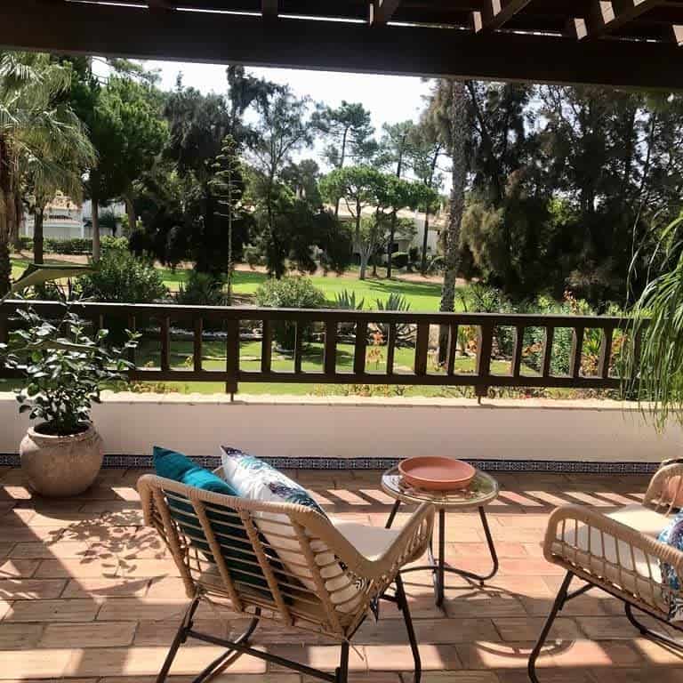 Sunny patio with rattan chairs, decorative pillows, small table, and a peaceful garden view.