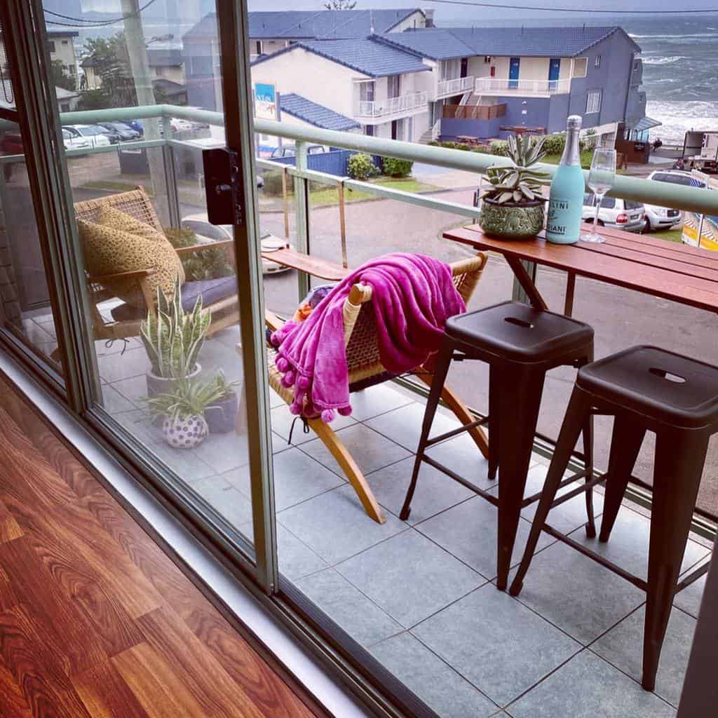 Cozy balcony with bar stools, a small table, a wicker chair, and potted plants overlooking the street.