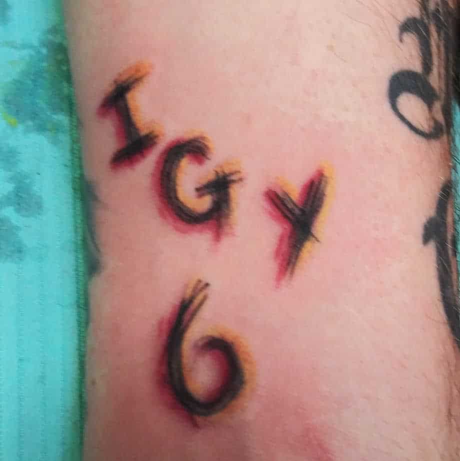 Forearm tattoo of 'IGY6' in bold, brushstroke-style lettering with red and yellow accents for a fiery effect