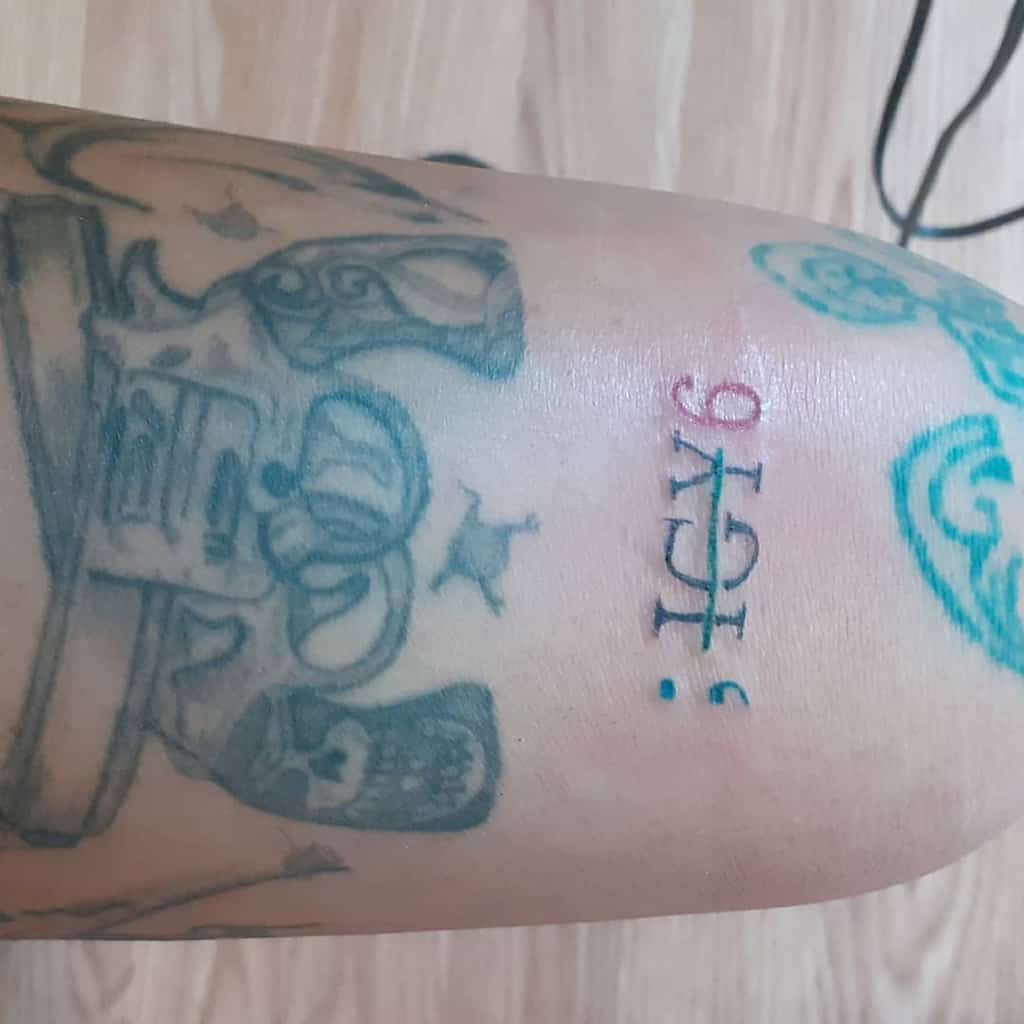 Minimalist ';IGY6' tattoo in black, blue, and red ink on the arm, placed among other tattoos with intricate designs