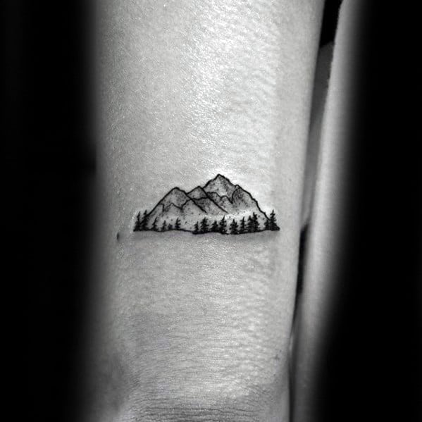 Minimalist mountain tattoo on the arm, featuring fine line peaks with subtle shading and a row of pine trees for a serene nature-inspired design.