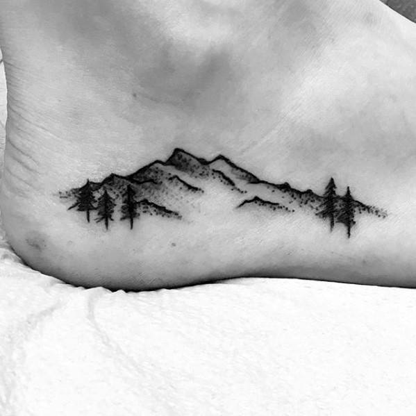 Minimalist mountain tattoo on the foot, featuring fine line peaks, subtle dot shading, and pine trees for a natural and artistic landscape design.