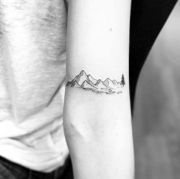 Minimalist mountain tattoo on the forearm, featuring fine line peaks, a lake, and a single tree for a serene and nature-inspired design.
